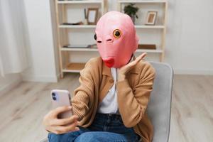 Lycklig rolig blond lady i rosa fisk mask värma Tröja håller på med selfie video ring upp Sammanträde i fåtölj på modern Hem interiör. paus från arbete, ta en ha sönder, social media i fri tid begrepp. kopia Plats foto