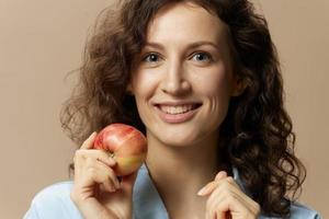 närbild porträtt av glad lockigt söt lady i jeans tillfällig skjorta med äpple nära kind Framställ isolerat på beige pastell bakgrund. friska mat. naturlig miljövänlig Produkter begrepp. kopia Plats foto