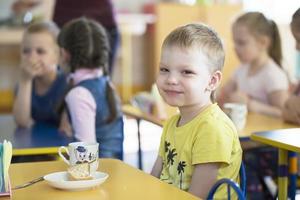 barn äta i dagis. pojke med en kopp på de tabell på de bakgrund av barn foto