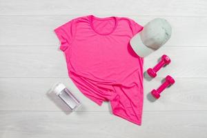rosa kvinna sport springa kondition t skjorta attrapp platt lägga. trä- bakgrund. sommar pilates sträckning tillbehör. sportig vatten flaska, hantlar, hatt. topp främre se t-shirt. mall tom kopia Plats foto