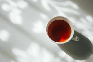 vit kopp av te på abstrakt bakgrund av naturlig ridå skugga faller på vit tabell. foto