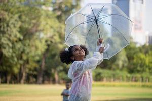 söt liten unge flicka spelar utomhus i de trädgård, barn flicka med paraply spelar regn i de parkera foto