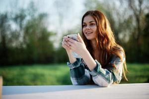 ung flicka bloggare frilansare innehav henne telefon ser på den och arbetssätt utomhus i en grön parkera eftertänksamt ser på de skärm uppkopplad foto