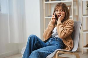 upphetsad Lycklig överraskad ung blond lady i värma Tröja skaffa sig Bra Nyheter från ring upp använda sig av telefon Sammanträde i fåtölj på Hem. paus från arbete, ta en ha sönder, social media i fri tid begrepp. kopia Plats foto