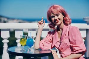 glad kvinna i en rosa klänning sitter på de terrass och drycker cocktails foto