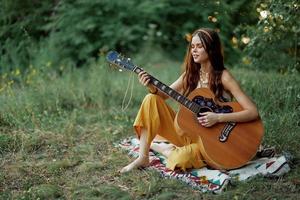 ung hippie kvinna Sammanträde utomhus på en pläd och spelar henne gitarr på de flodbank in i de solnedgång foto