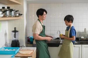Lycklig leende ung asiatisk far och son tvättning maträtter i kök på Hem foto