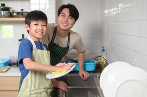 Lycklig leende ung asiatisk far och son tvättning maträtter i kök på Hem foto