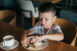 liten och söt pojke har frukost i Kafé. äter pannkakor med sur grädde foto