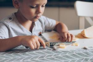 söt pojke 4-5 år gammal med kniv skärande en ost på de skärande styrelse på de tabell i de kök foto
