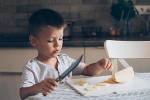 söt pojke 4-5 år gammal med kniv skärande en ost på de skärande styrelse på de tabell i de kök foto