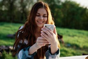 en skön kvinna avkopplande och arbetssätt på henne telefon Sammanträde i natur i de parkera bland de träd leende och innehav henne smartphone i henne hand belyst förbi de ljus solnedgång ljus foto