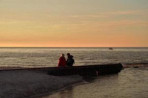 landskap med solnedgång på de baltic hav med en vågbrytare och en par av människor Sammanträde på den foto
