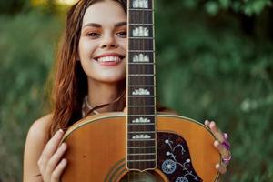 ung hippie kvinna med eco bild leende och ser in i de kamera med gitarr i hand i natur på en resa foto