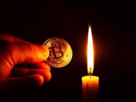 guld bitcoin mynt i handen i det varma ljuset av ett ljus på en mörk bakgrund foto