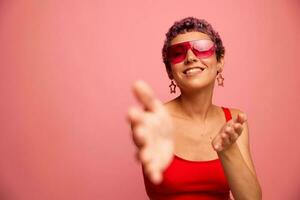 mode porträtt av en kvinna med en kort frisyr i färgad solglasögon med ovanlig Tillbehör med örhängen ler på en rosa ljus bakgrund visar henne händer till de kamera foto