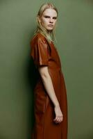 självsäker modern blond ung modell ser över henne axel på kamera klänning i brun läder dike täcka Framställ isolerat på över oliv grön studio vägg bakgrund. eleganta erbjudande foto