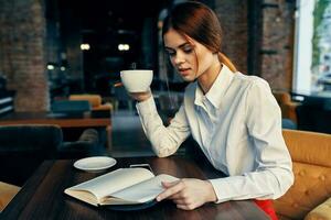 kvinna i kjol skjorta på tabell i Kafé kaffe kopp i hand och anteckningsblock penna foto