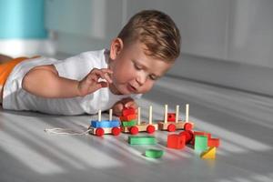 bebis lägger ner en tåg från geometrisk block. de pojke 2 år lögner på de golv och spelar med en färgrik trä- stapling tåg för barn. foto