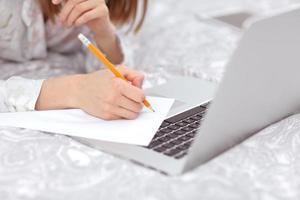 närbild av en kvinnas hand med en penna för att skriva. flicka som arbetar, lär sig och använder bärbar dator i sovrummet. frilansare. skriva, skriva. kommunikation och teknik, självstudiekoncept. foto