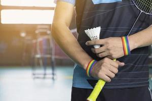 badminton spelare bär regnbåge armband och innehav racket och vit fjäderboll i främre av de netto innan tjänande den till spelare i annan sida av de domstol, begrepp för HBTQ människor aktiviteter. foto