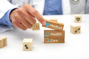 hand av läkare innehav en trä- blockera kub med text och Bakom den är en trä- blockera med suddig ikon symbol. medicinsk och hälsa begrepp. foto
