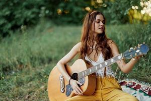 flicka hippie kvinna spelar gitarr i miljövänlig kläder Sammanträde på de jord utanför i natur i de falla tittar på de solnedgång foto
