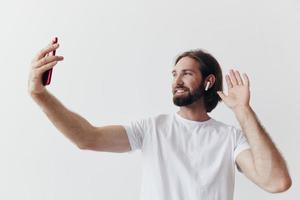 en manlig bloggare videoinspelning han själv på hans telefon och chattar med människor uppkopplad med en leende i en vit t-shirt mot en vit vägg foto