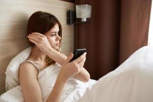 kvinna lögner i säng under de omslag med en mobil telefon i henne hand interiör gardiner design foto