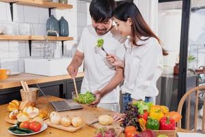 ung asiatisk par matlagning med frukt och grönsaker och använder sig av bärbar dator i de kök till laga mat mat tillsammans inom de familj lyckligt, familj begrepp. foto