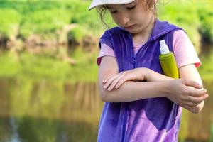 flicka sprayar myggspray på huden i naturen som biter hennes händer och fötter. skydd mot insektsbett, avstötande säkert för barn. friluftsliv, mot allergier. sommartid foto