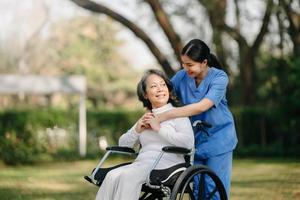 äldre asiatisk senior kvinna på rullstol med asiatisk försiktig vårdgivare och uppmuntra patient, gående i trädgård. med vård från en vårdgivare och senior hälsa försäkring. foto