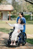 äldre asiatisk senior kvinna på rullstol med asiatisk försiktig vårdgivare och uppmuntra patient, gående i trädgård. med vård från en vårdgivare och senior hälsa försäkring. foto