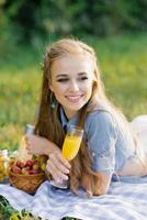 Söt kvinna med en glas av juice i henne hand är liggande på en filt på en sommar picknick foto