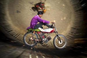 bangladesh februari 14, 2018 en lantlig hämma cyklist är riskerar hans liv förbi skalning de vägg av en stor trä- väl i en motorcykel karneval sidoshow på dohar, dhaka. foto