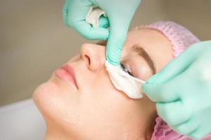 ung kvinna tar emot ögonhår avlägsnande procedur och tar bort mascara med en bomull svabb och pinne i en skönhet salong. foto