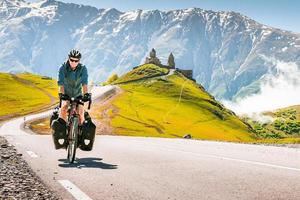 cyklist tillbaka se på de väg i naturskön kaukasus natur med gergeti trinity kloster i de bakgrund. resande på cykel. solo- resa lång distans cykel touring begrepp foto