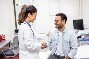 leende ung kvinna läkare skaka hand stänga hälsa försäkring handla med äldre patient på samråd i sjukhus. Lycklig kvinna gp handslag hälsning skaffa sig bekant med man i klinik. foto