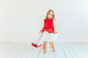 ljuv liten flicka i röd klänning Sammanträde på stol mot vit vägg på Hem, avkopplande i vit ljus levande rum inomhus. barndom skolbarn ungdom koppla av begrepp. foto