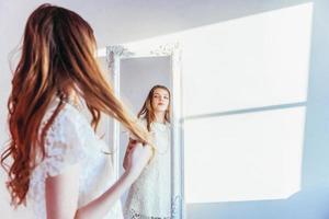 skönhet smink morgon- rutin kärlek själv begrepp. ung Tonårs flicka ser på reflexion i spegel. ung positiv kvinna bär vit klänning Framställ i ljus ljus rum mot vit vägg. foto