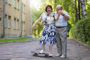 Lycklig äldre par med en skateboard. stilig man och kvinna senior medborgare. Make och fru av gammal ålder för en promenad i de stad. foto