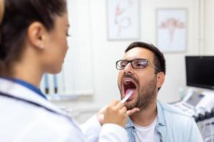 manlig patient öppning hans mun för de läkare till se på hans hals. kvinna läkare granskning öm hals av patient i klinik. otolaryngologist undersöker öm hals av patient. foto