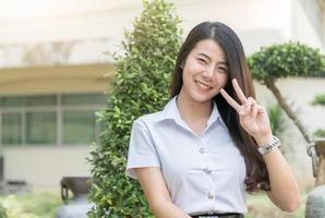 söt ung asiatisk kvinna i enhetlig studerande visa v tecken finber, leende och ser på kamera, Lycklig och koppla av begrepp foto