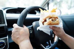 asiatisk kvinna förare håll och äta hamburgare och franska frites i bil, farlig och risk ett olycka. foto