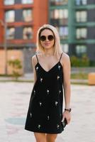 Lycklig leende blond kvinna i en svart klänning promenader i de stad i sommar foto
