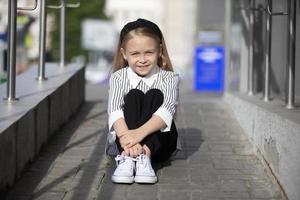 en liten flicka sitter på de trottoar i de gata. en barn i stad. foto