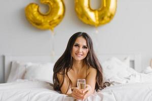 lång håriga födelsedag kvinna lögner på de säng i de morgon- och innehar en glas av champagne i henne händer och ler fira henne trettionde födelsedag foto