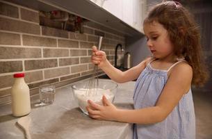 förtjusande europeisk liten flicka i blå klänning blandning mjöl med torr Ingredienser i en glas skål använder sig av en vispa, framställning pannkaka deg i de Hem kök för skrava tisdag foto