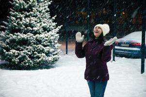 glad kvinna i värma kläder, gläds i de först snö, stående på en snöig gata upplyst förbi ny år kransar foto