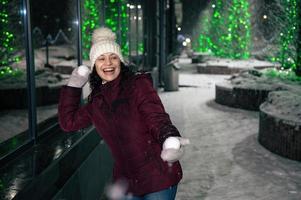 charmig kvinna har roligt i en stad gata, upplyst förbi girlander, spelar med snöbollar på en vinter- snöig kväll foto